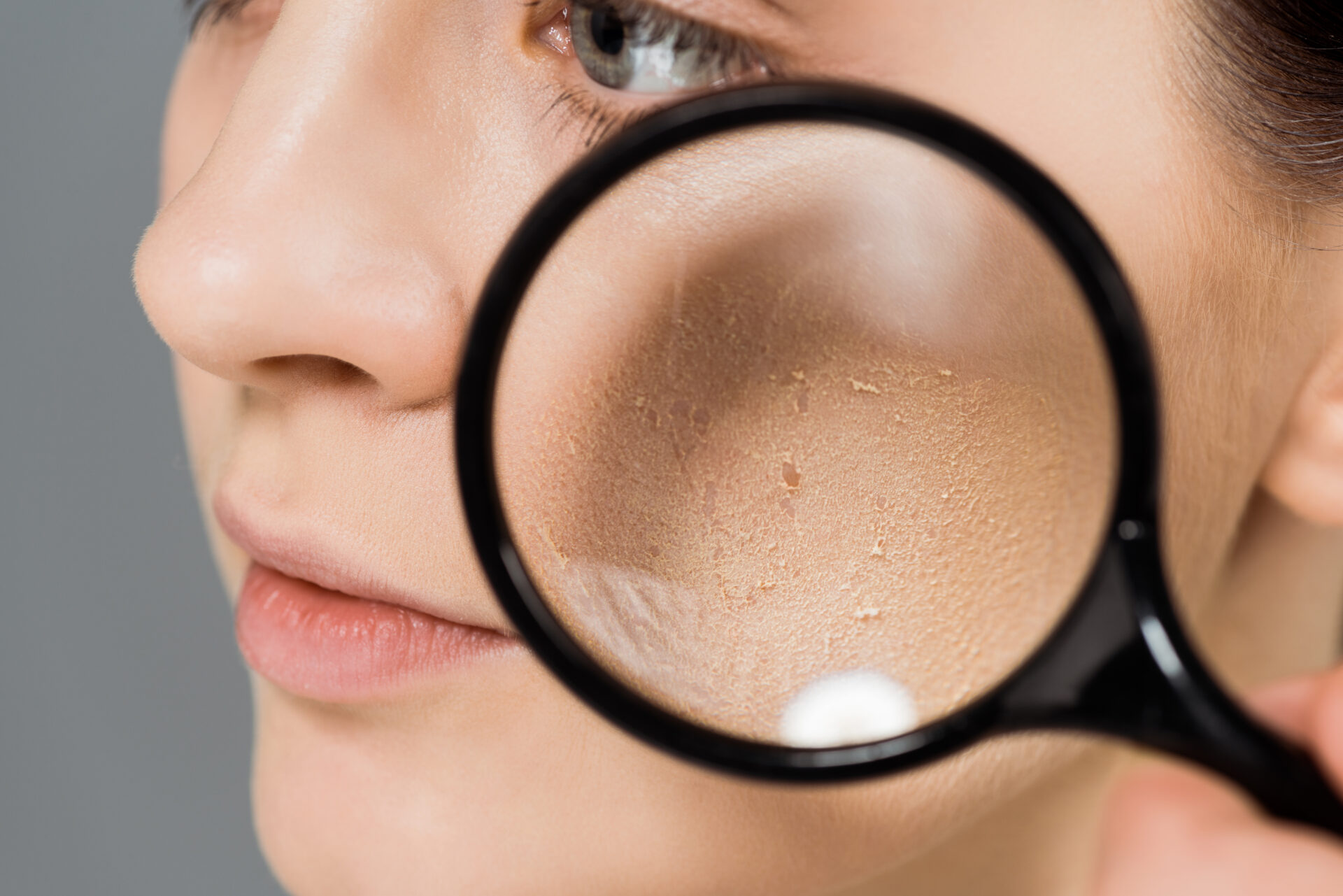 cropped view of young woman with magnifier near problem skin isolated on grey